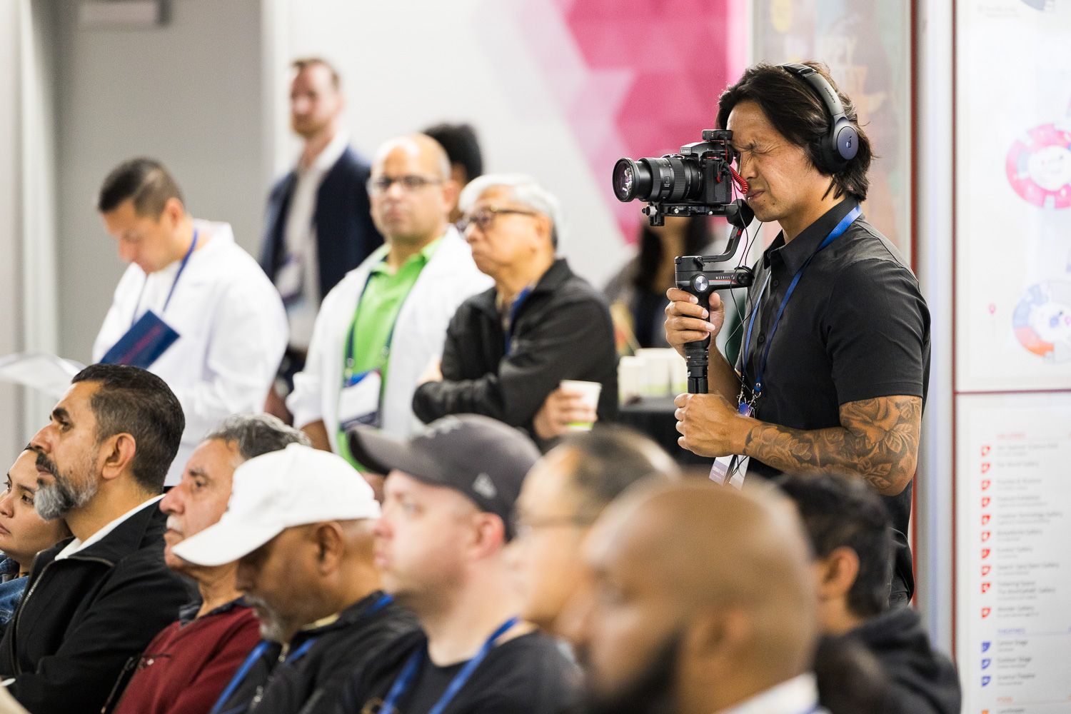 Videographer with camera on gimbal films audience at indoor event while attendees sit in rows listening attentively.