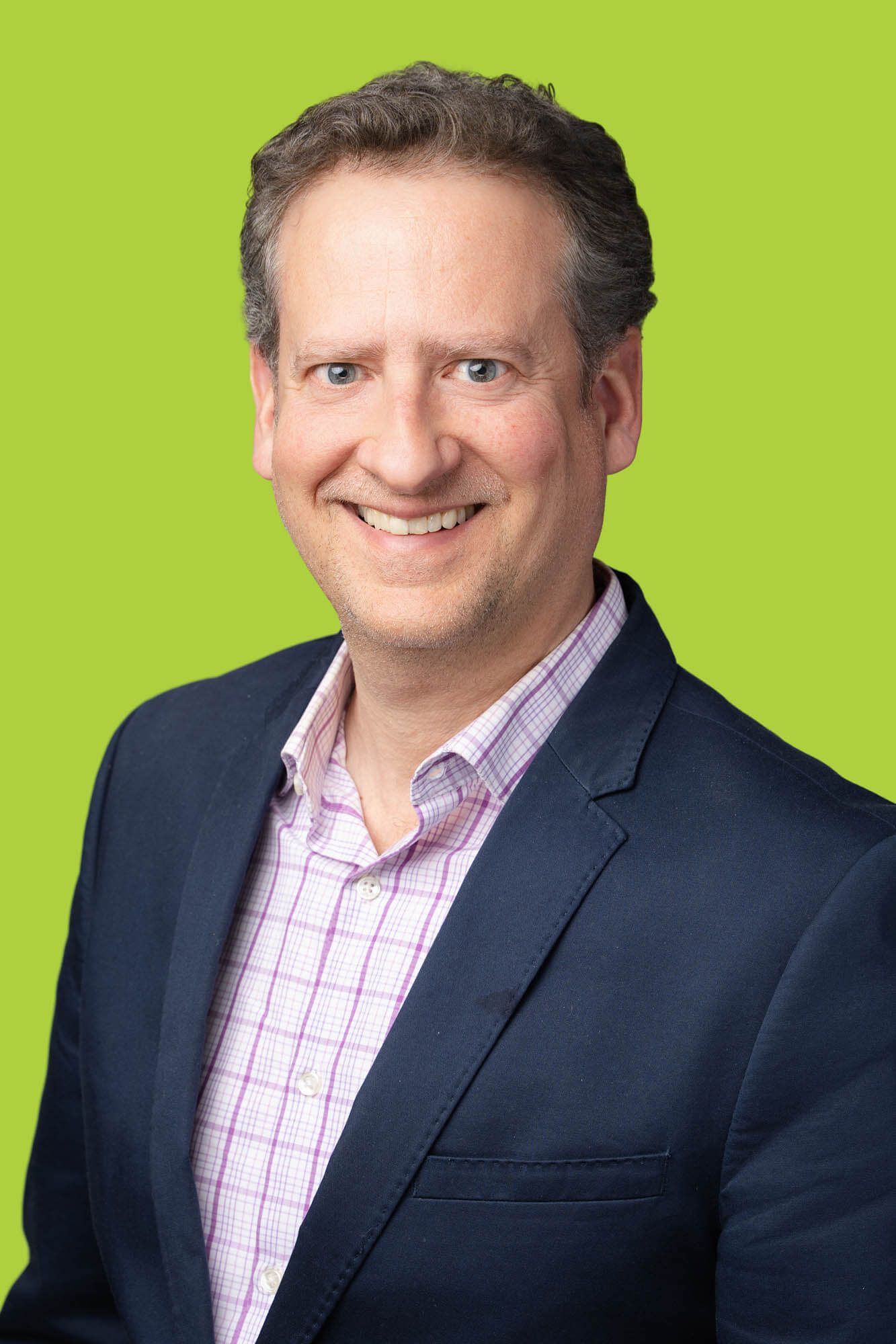 Professional headshot of a smiling middle-aged man in a navy blazer and pink plaid shirt against a bright green background.