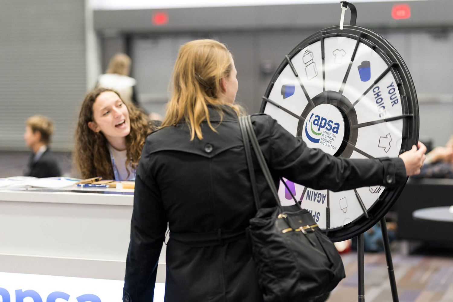 Woman spins APSA prize wheel at a conference booth while a smiling representative looks on.