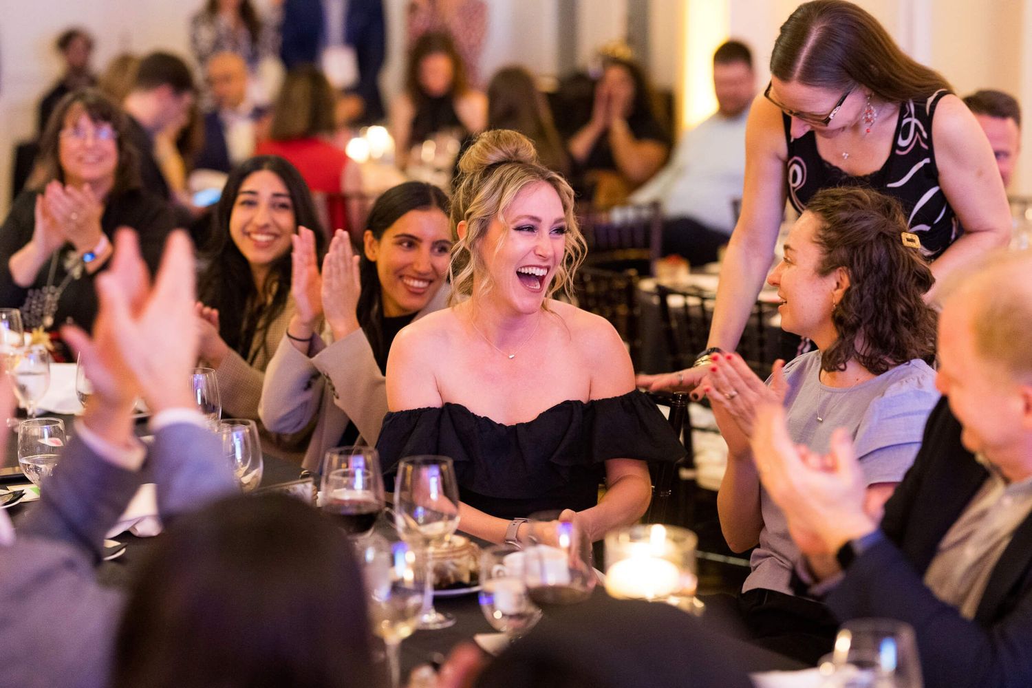 Woman in black off-shoulder dress laughing joyfully at a formal dinner event with applauding guests