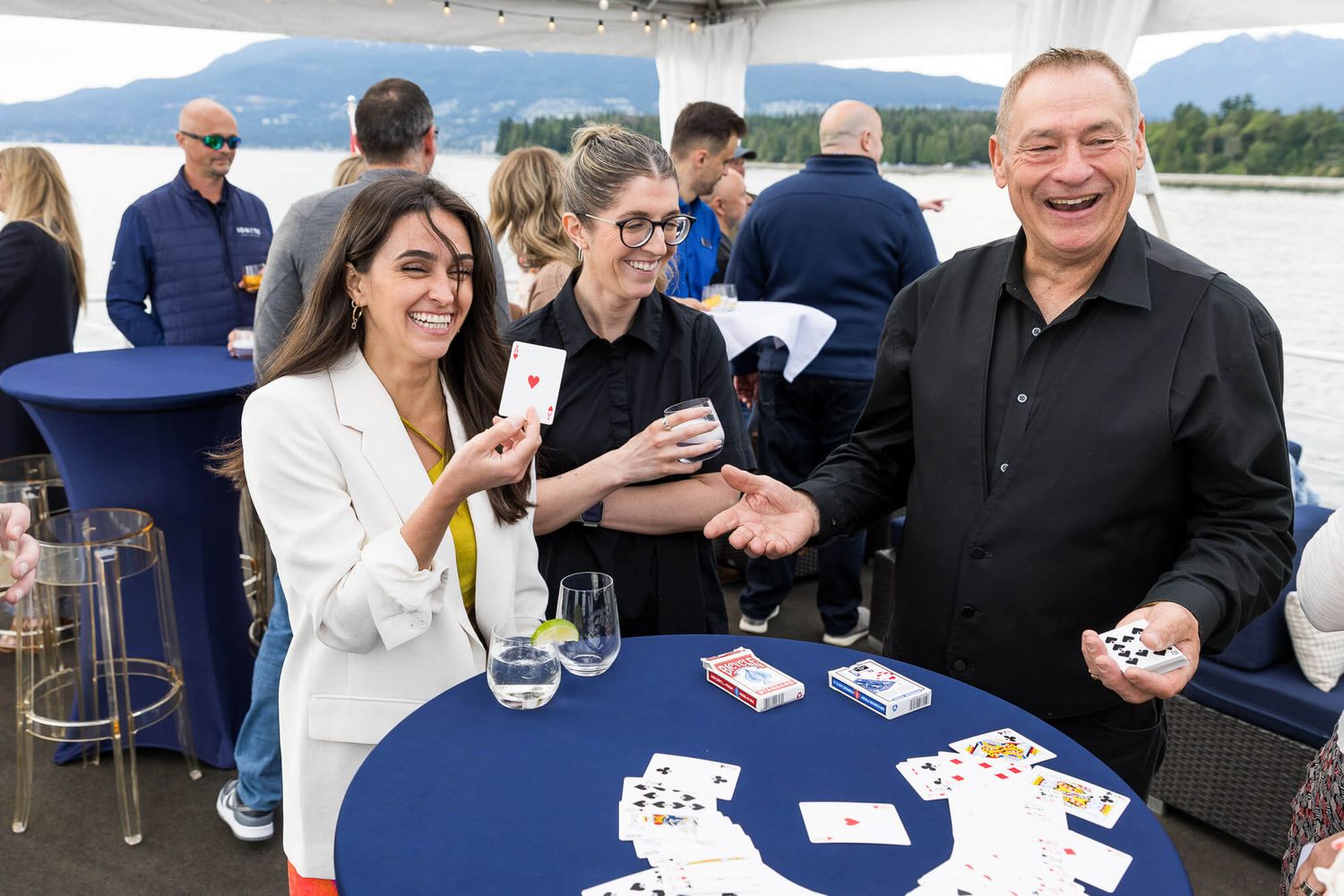 Three people smile and laugh while playing cards at a blue high-top table during an outdoor waterfront event.