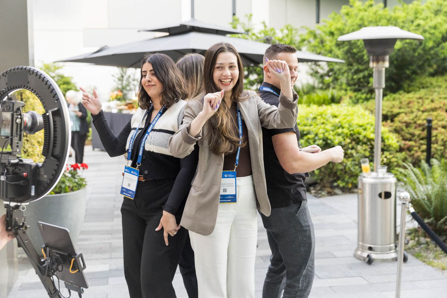 Three professionals in lanyards pose playfully outdoors at a corporate event near a ring light and patio heater.