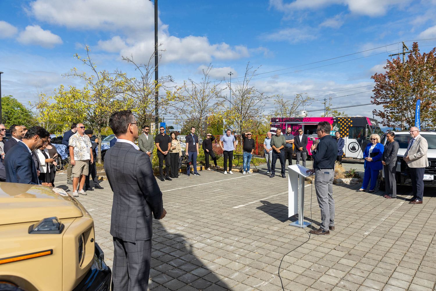 Outdoor public speaking event with a crowd gathered around a podium in a sunny parking lot with trees and vehicles nearby.