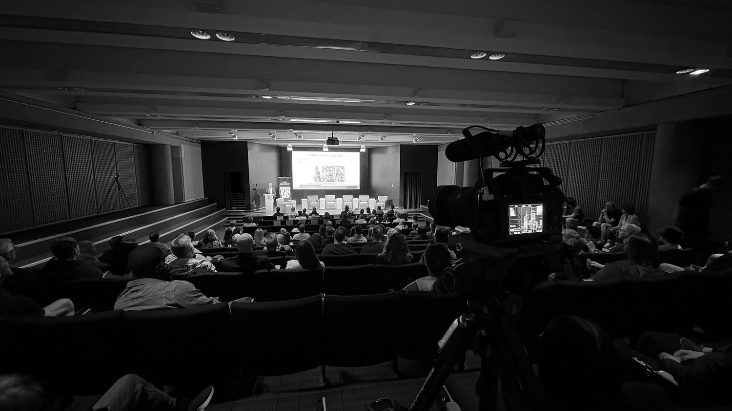 Black and white photo of a conference auditorium with audience, panel on stage, and a video camera in the foreground.