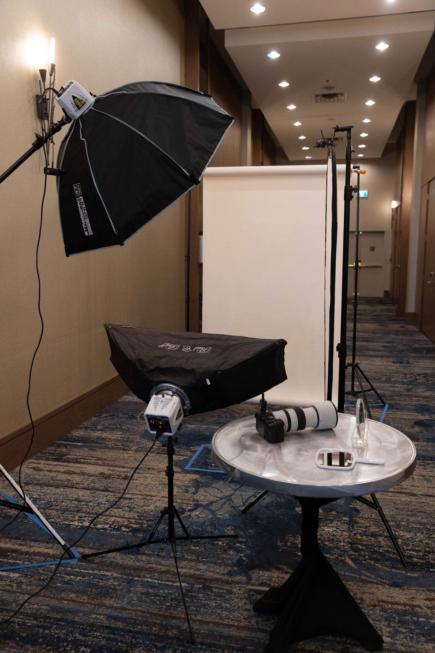 Professional photography studio setup with softbox umbrella lights, white backdrop, and camera on a round table in a hotel hallway.