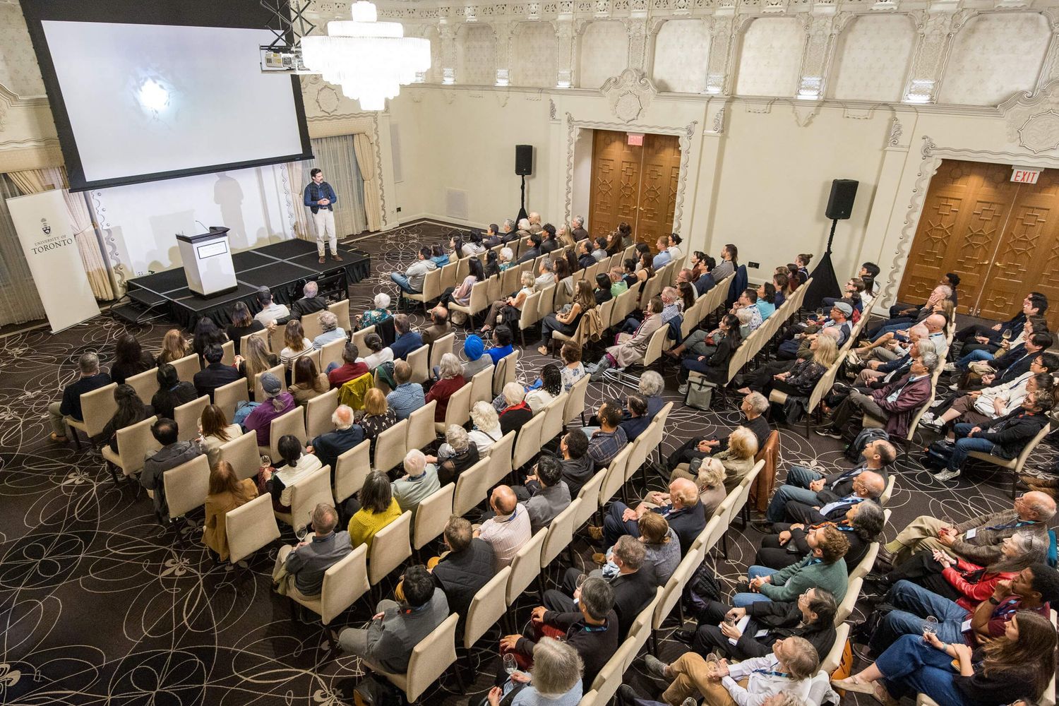 Hall filled with attendees seated in rows, listening to a speaker presenting on stage with a projection screen located at Rosewood Hotel