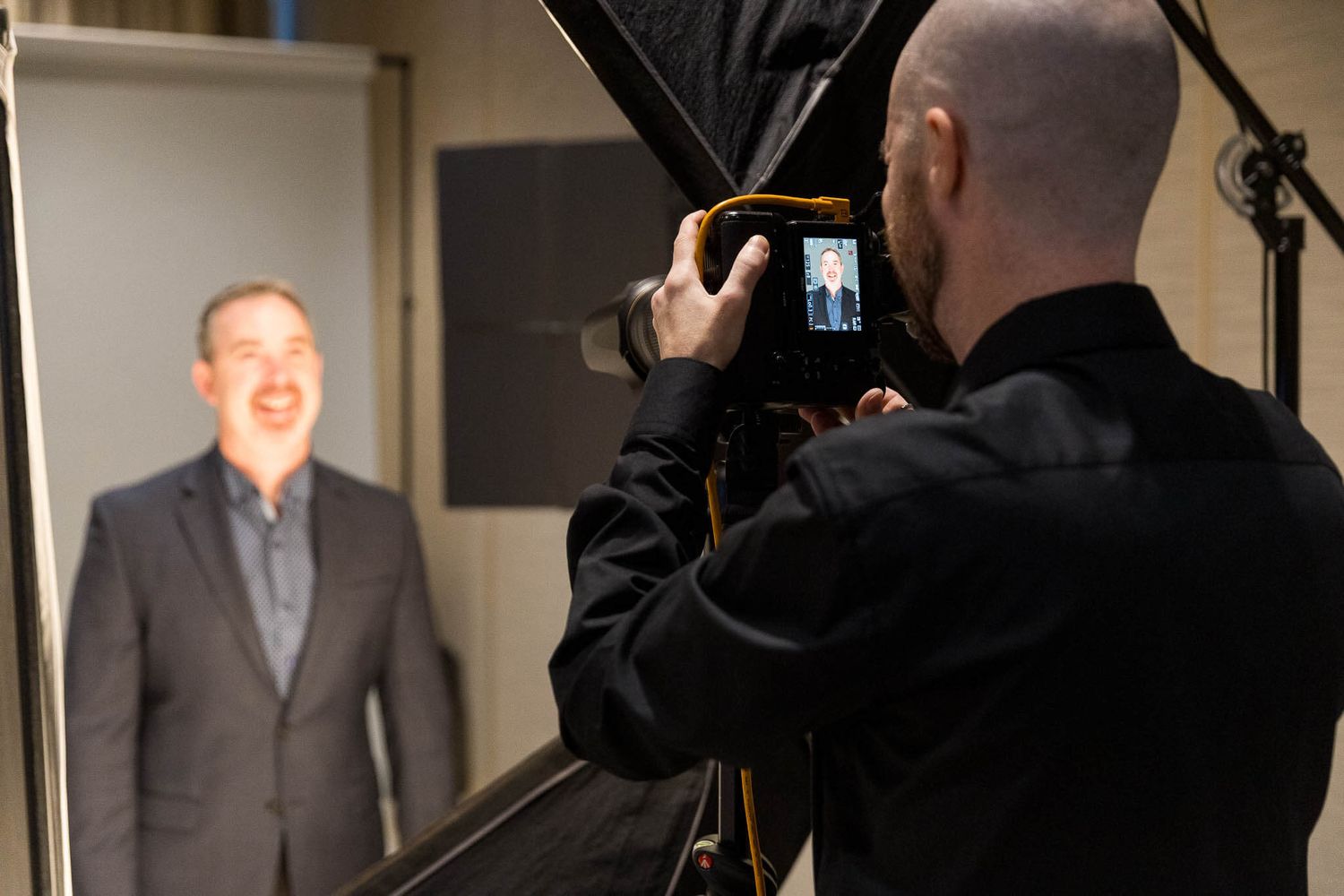 Photographer capturing a professional headshot of a smiling man in a suit using a camera with softbox lighting in a studio.