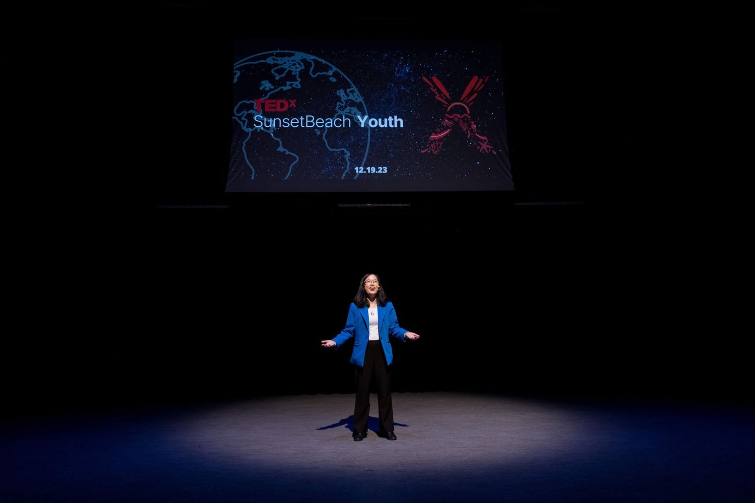 Speaker presenting on stage at TEDx SunsetBeach Youth event under spotlight with branded screen backdrop.