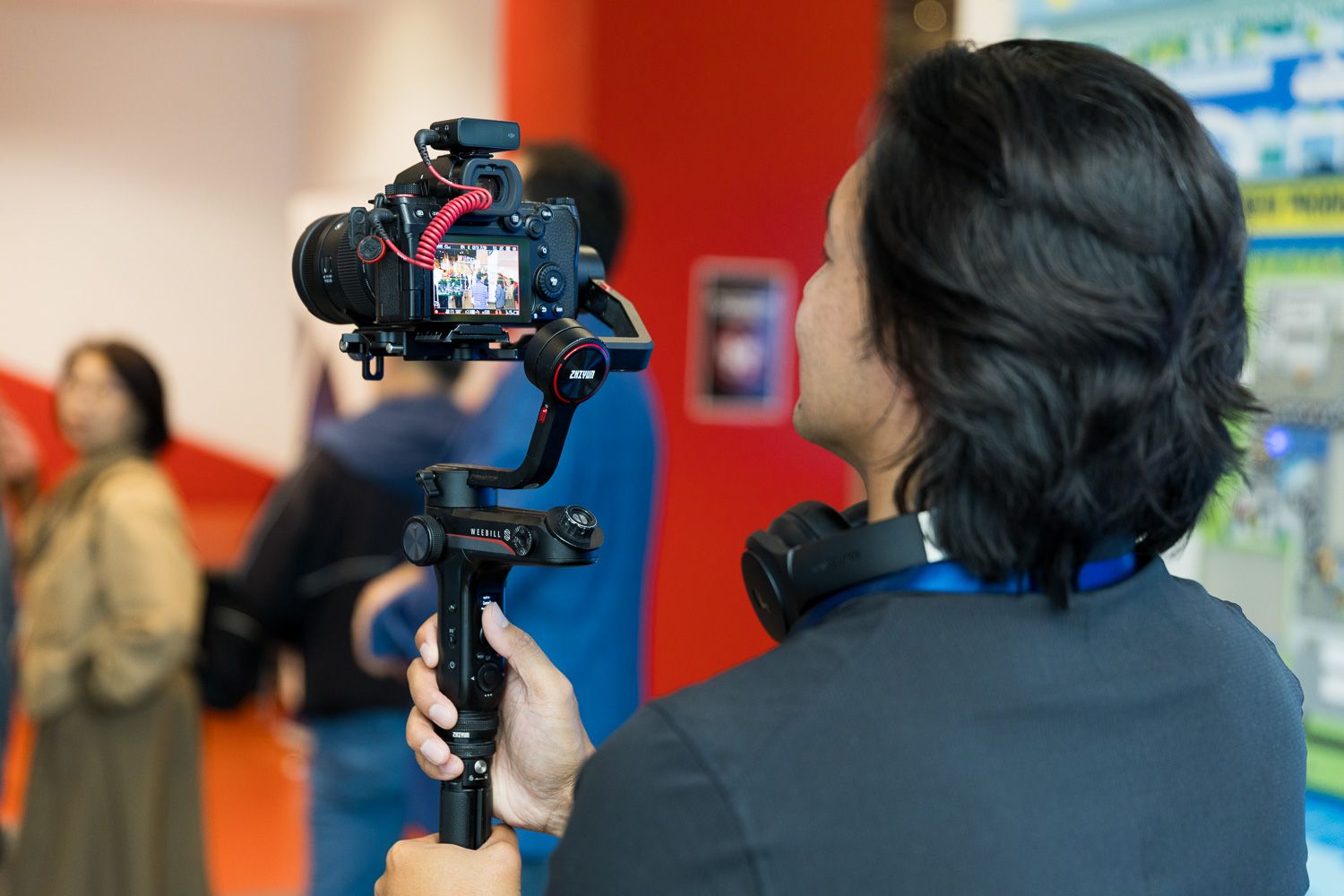 Videographer using a camera gimbal stabilizer to film an event indoors with people in the background.