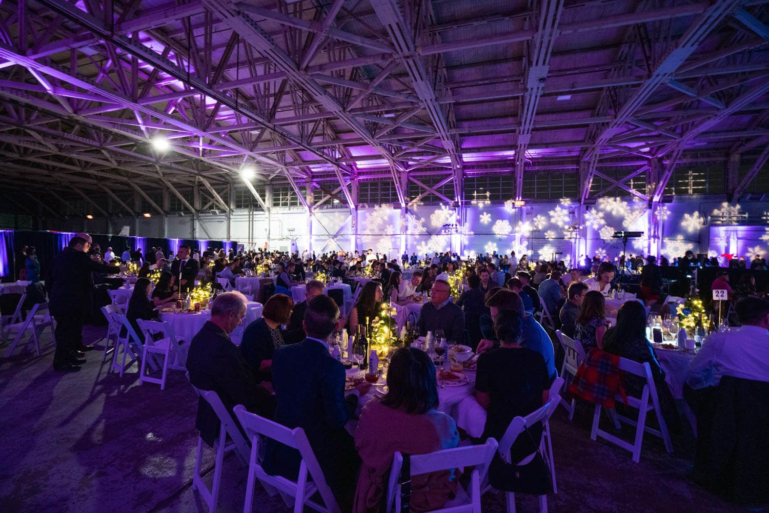 Elegant gala dinner event with guests seated at decorated tables under purple lights in a large venue.