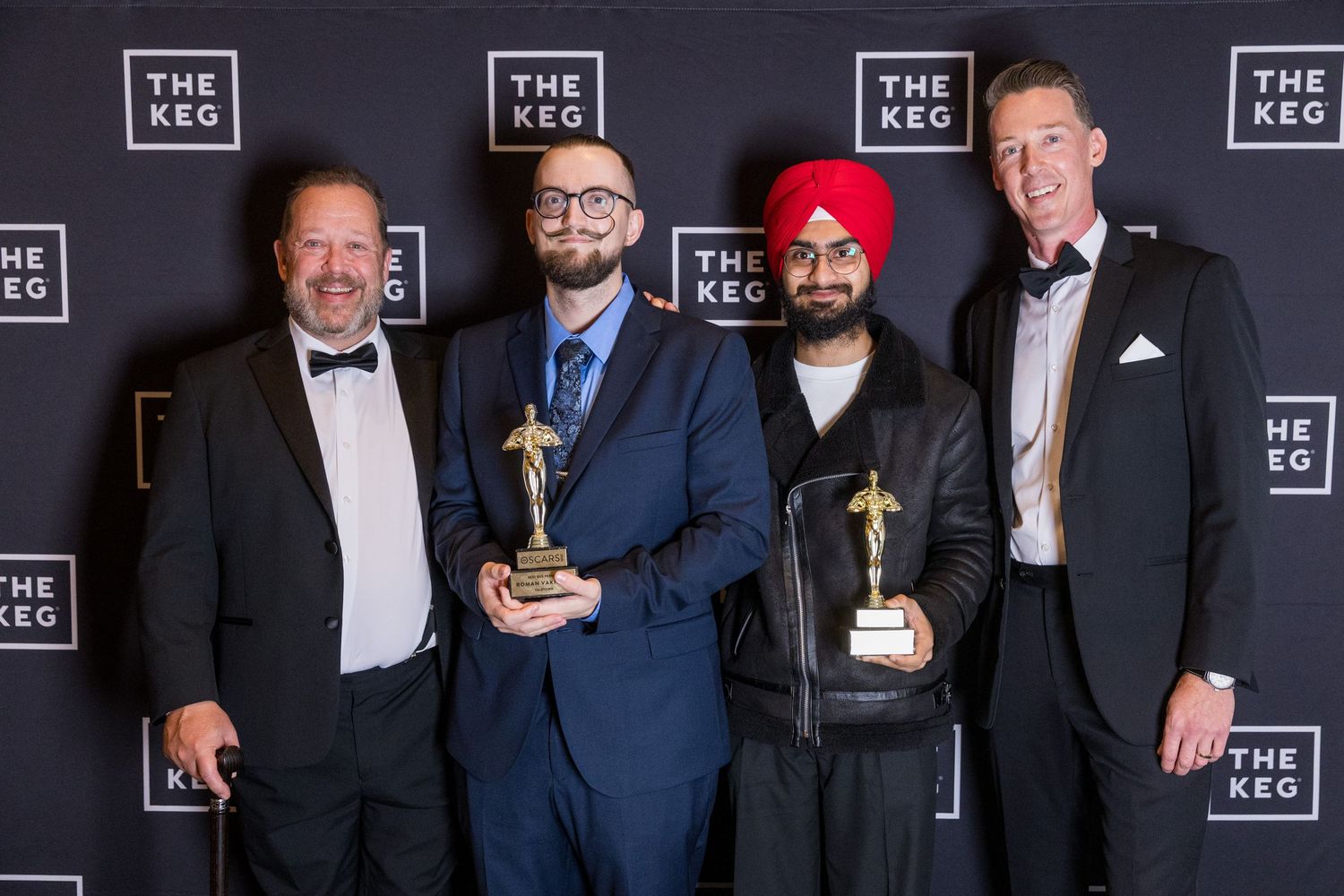 Four men in formal attire pose with trophy awards in front of The Keg restaurant branded backdrop.