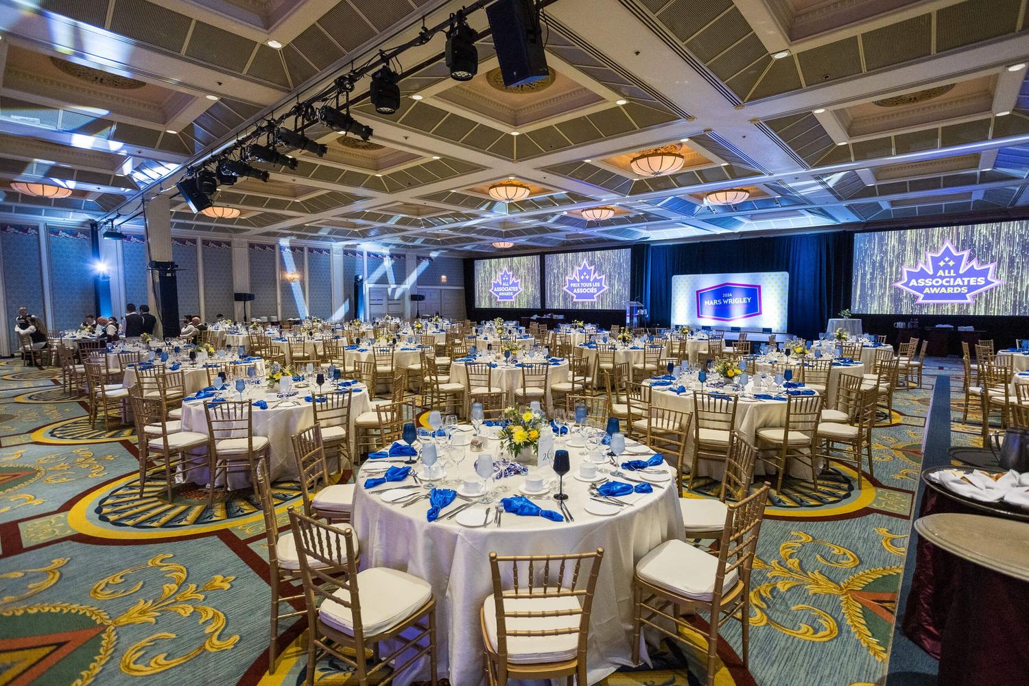 Elegant banquet hall with round tables, gold chairs, blue linens, and large projection screens for a formal event.