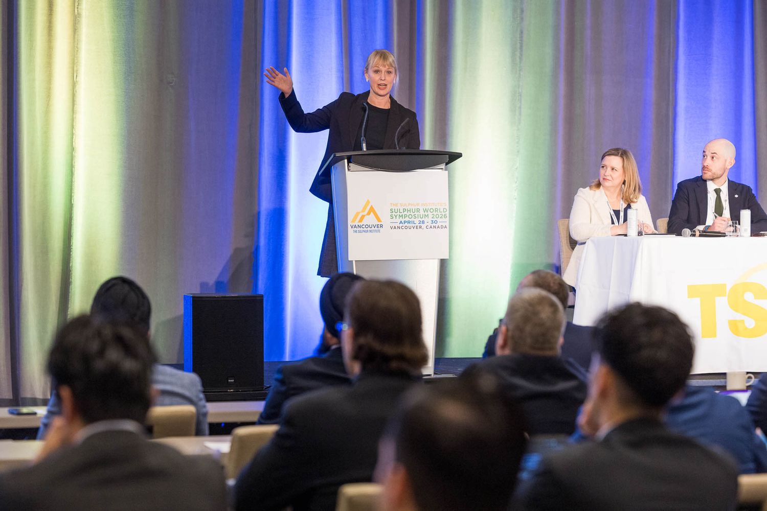 Speaker at podium addresses audience at professional conference, with two panelists seated at table on stage.