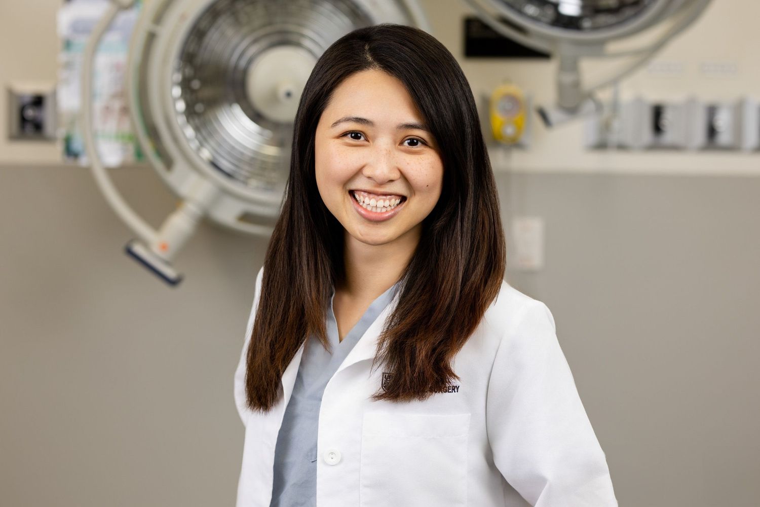 Smiling female doctor in white lab coat standing in medical facility with surgical light in background.