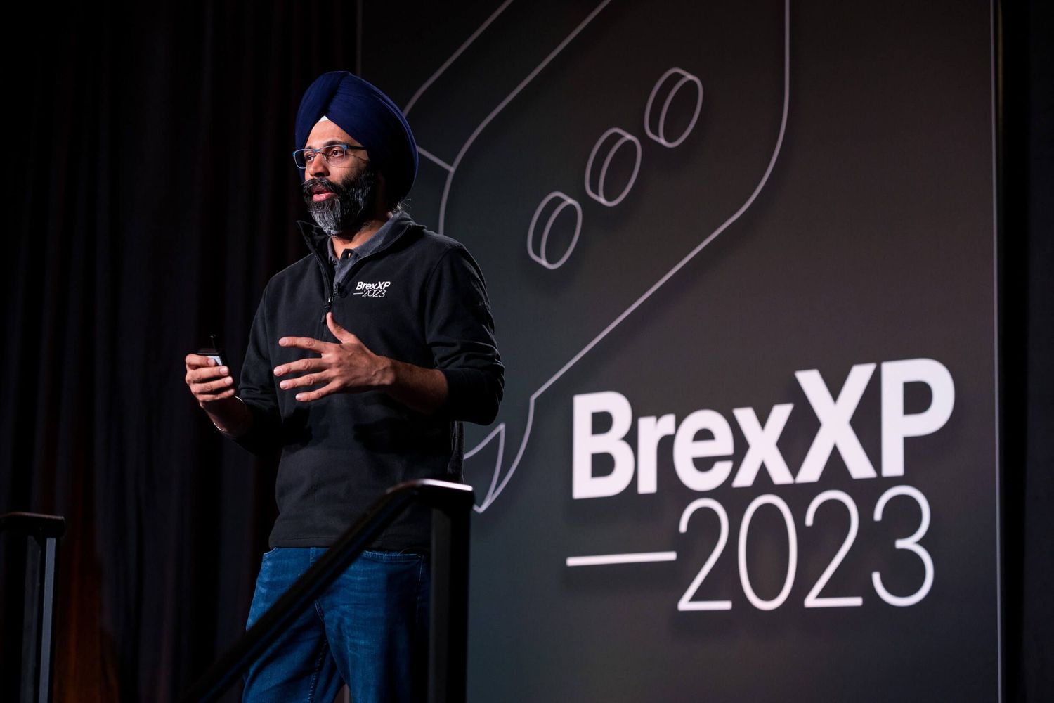 Man in blue turban speaks at BrexXP 2023 conference on dark stage with branded backdrop.