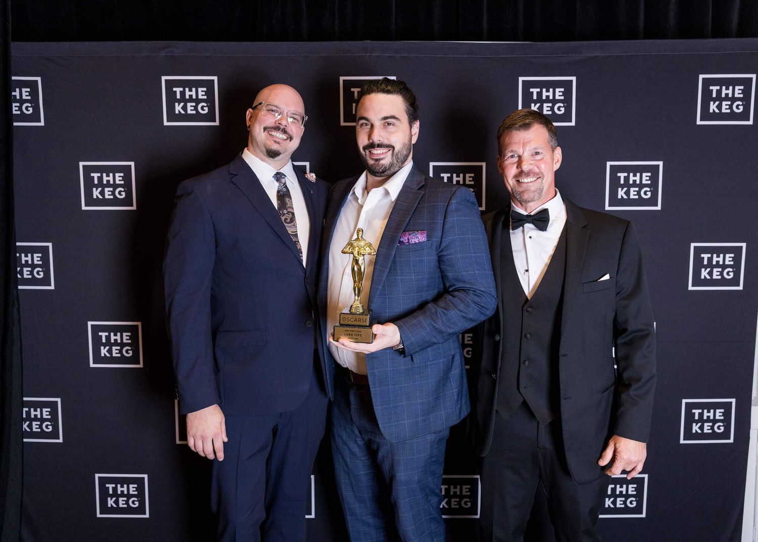 Three men in formal attire pose with a trophy at The Keg awards event backdrop.