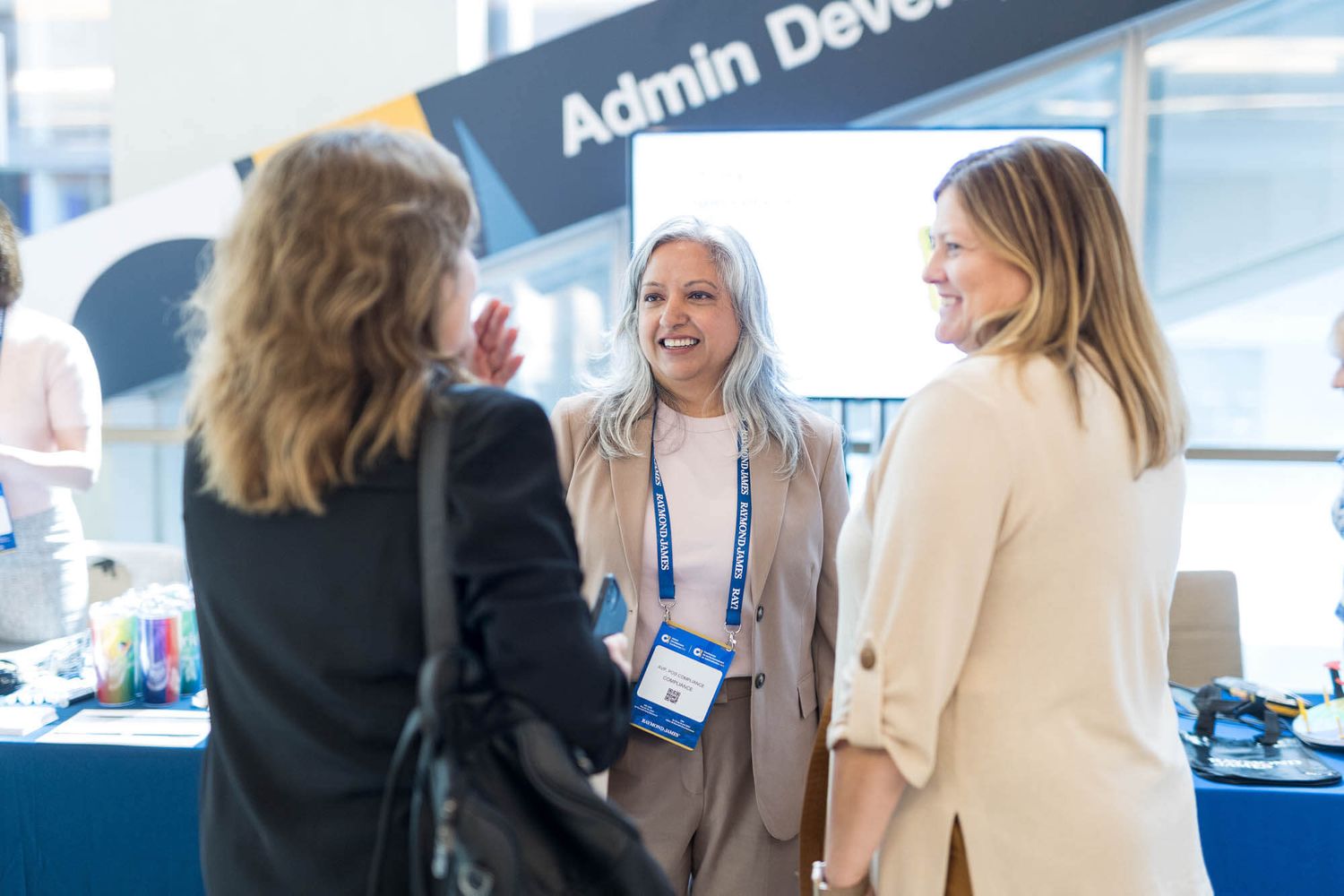 Three professional women networking and smiling at an Admin Dev conference booth event.