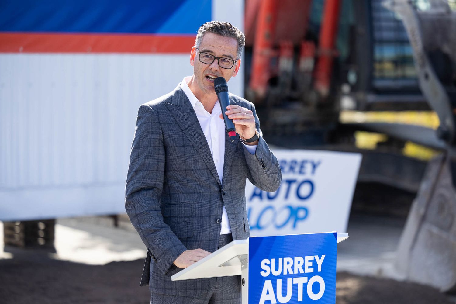 Man in suit speaking at Surrey Auto Loop event podium outdoors.
