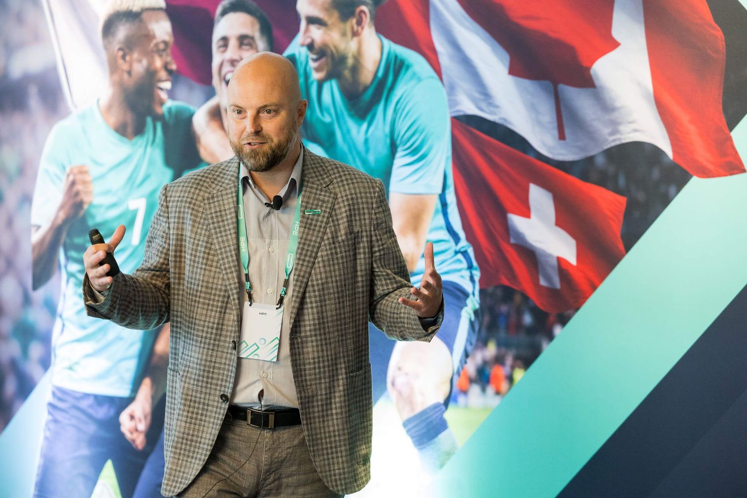 Male speaker in gray blazer presenting at sports corporate event with soccer players mural behind him.