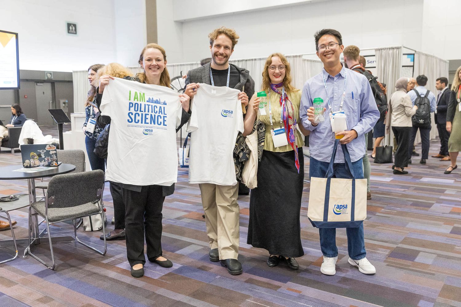 Four people smiling at a conference, one holding a Political Science t-shirt, with lanyards and tote bags visible.
