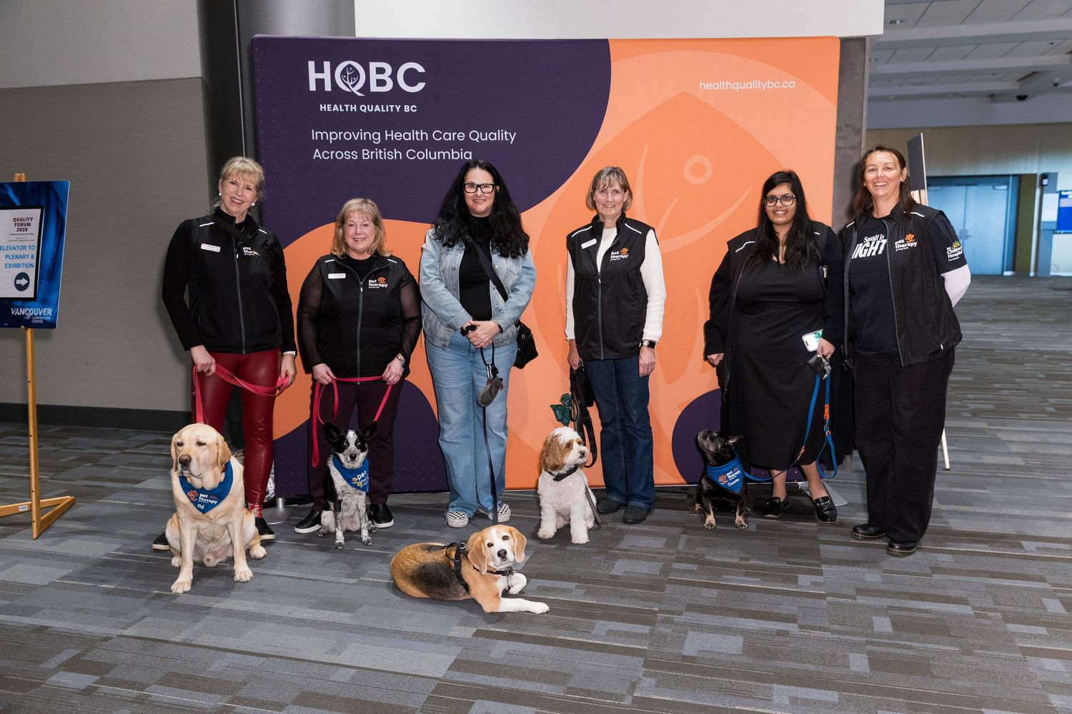 Six people with therapy dogs pose in front of a Health Care Quality banner at the BC Quality Forum held at the Vancouver Convention Centre.