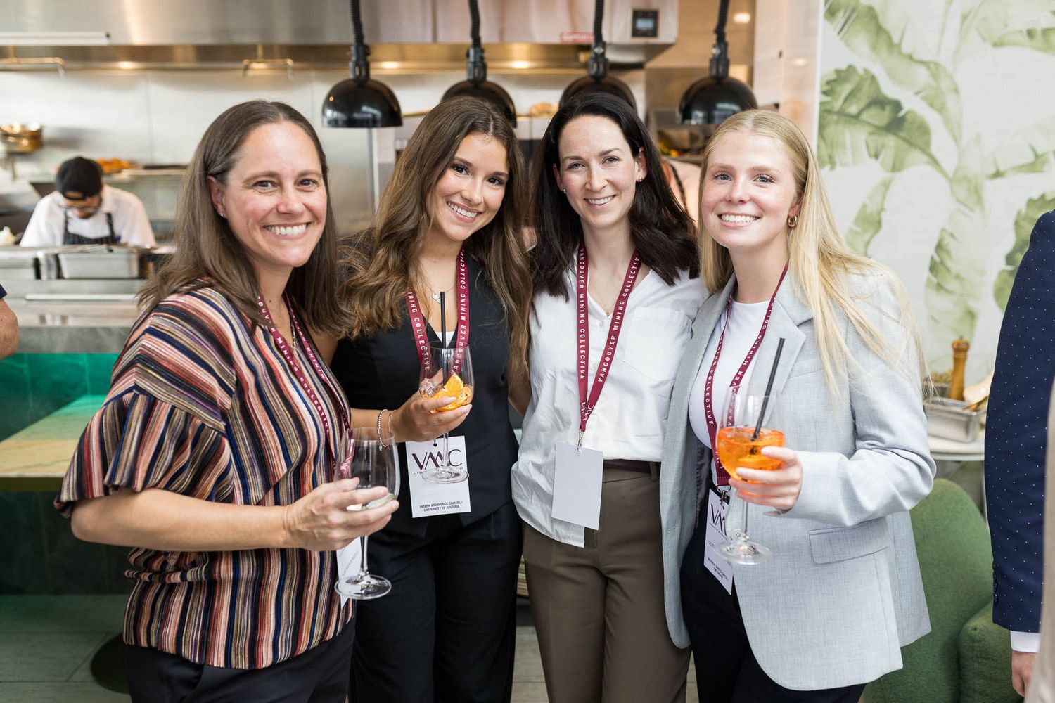 Four women smiling and holding drinks at a professional networking event indoors.