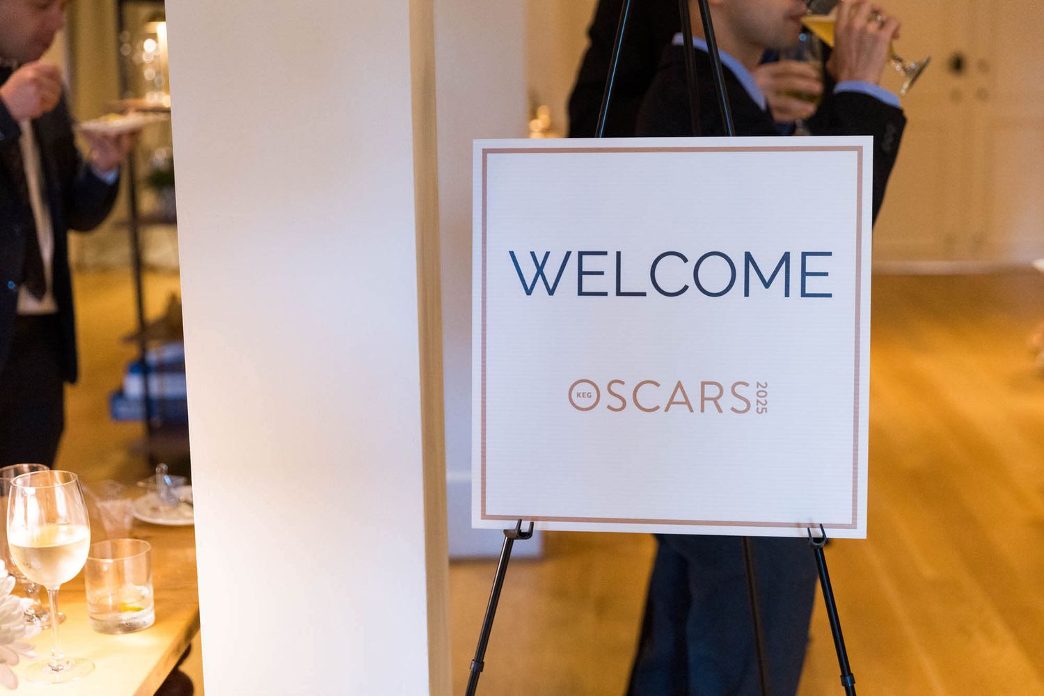 Welcome sign on easel for the Oscars event, with guests mingling in the background at an elegant venue.