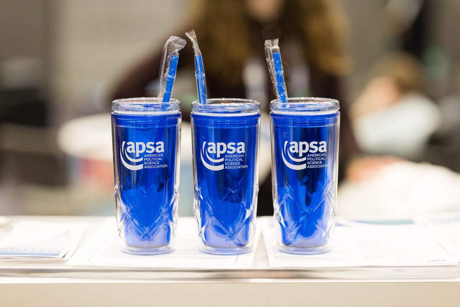 Three blue APSA branded tumbler cups with straws displayed on a table at a political science conference.