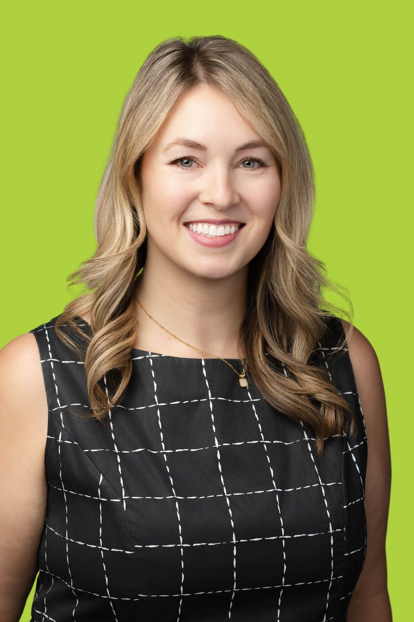 Smiling blonde woman in black grid-pattern dress poses for professional headshot on bright green background.
