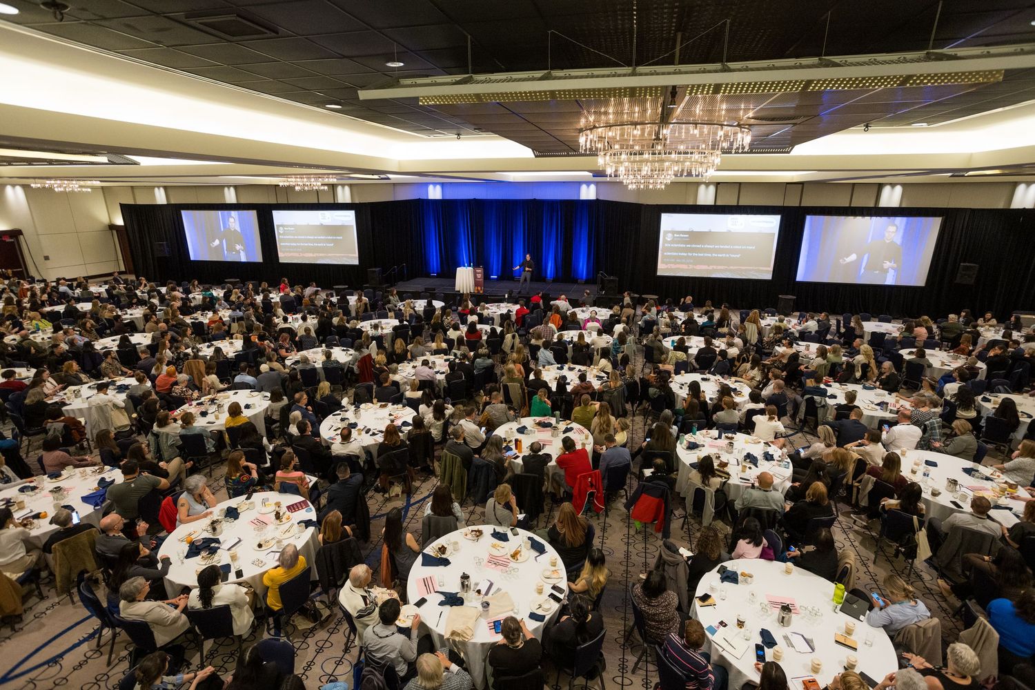 Large conference banquet hall filled with attendees seated at round tables facing presentation screens and a podium.