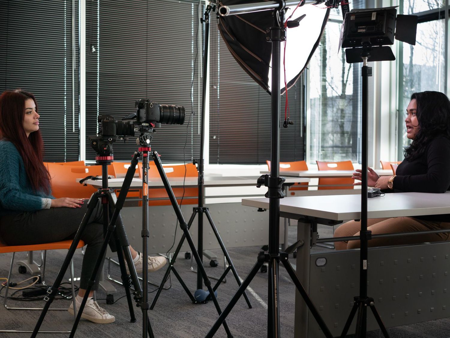 Two women in a professional video shoot setup with studio lights, camera on tripod, and orange chairs in modern office.