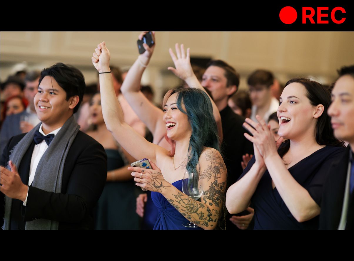 Excited crowd cheering and clapping at a live event, woman with tattoos raising hands in celebration.