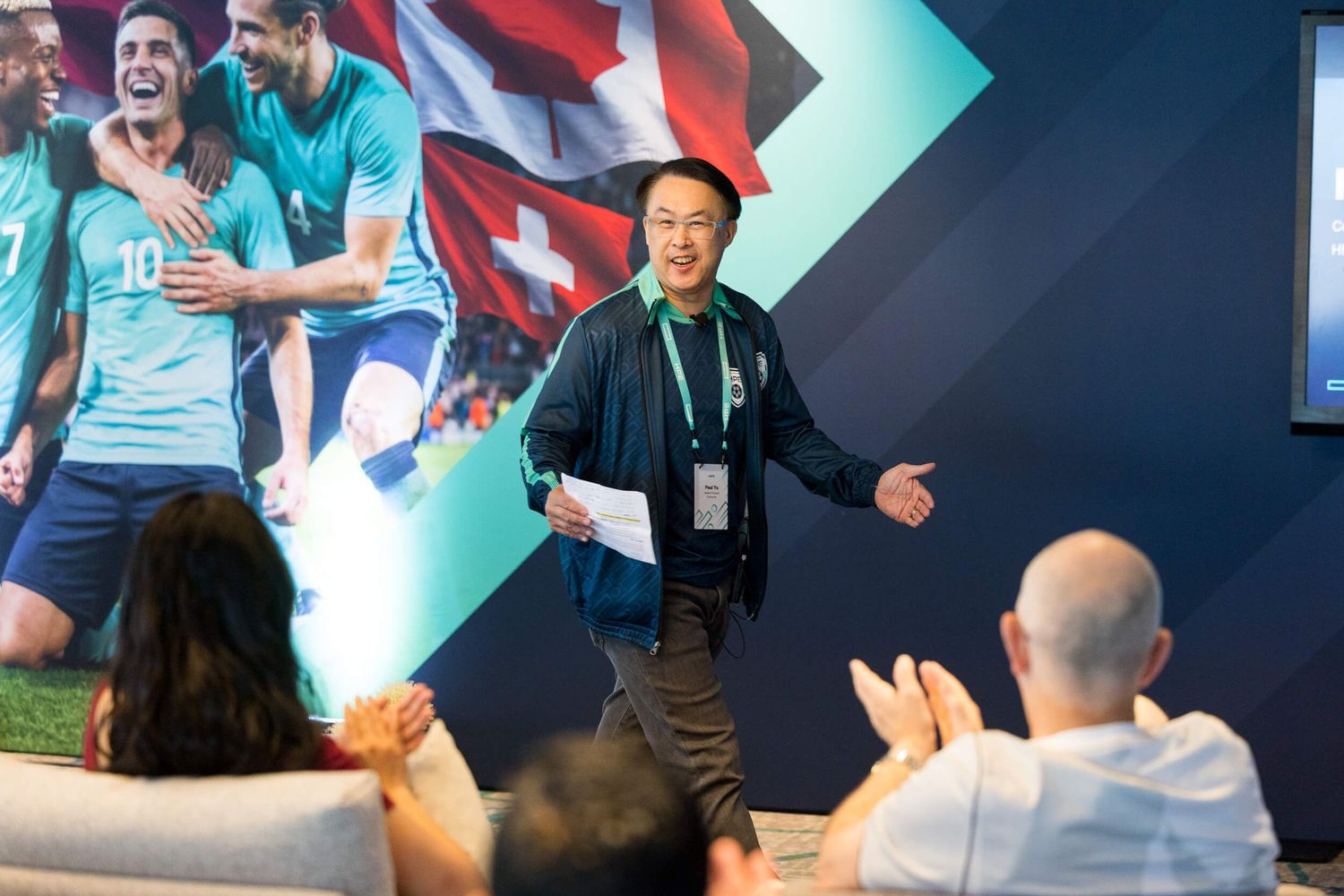 Asian male speaker presenting at sports-themed corporate event with soccer mural backdrop.