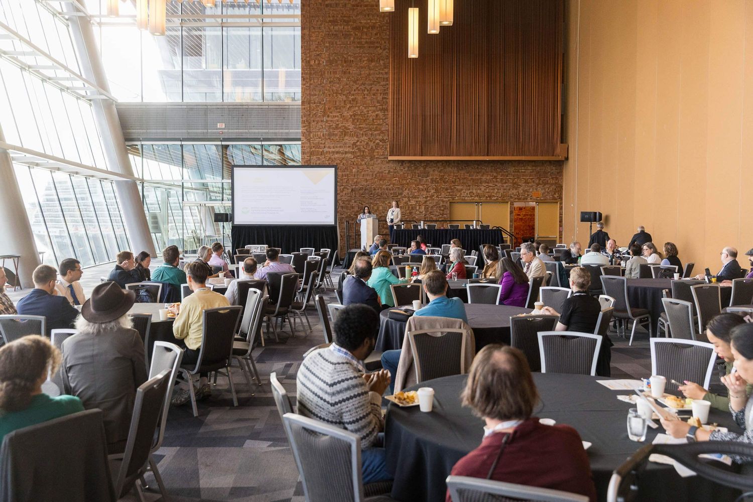 Attendees seated at round tables facing a presentation screen and speaker inside the Vancouver Convention Centre.