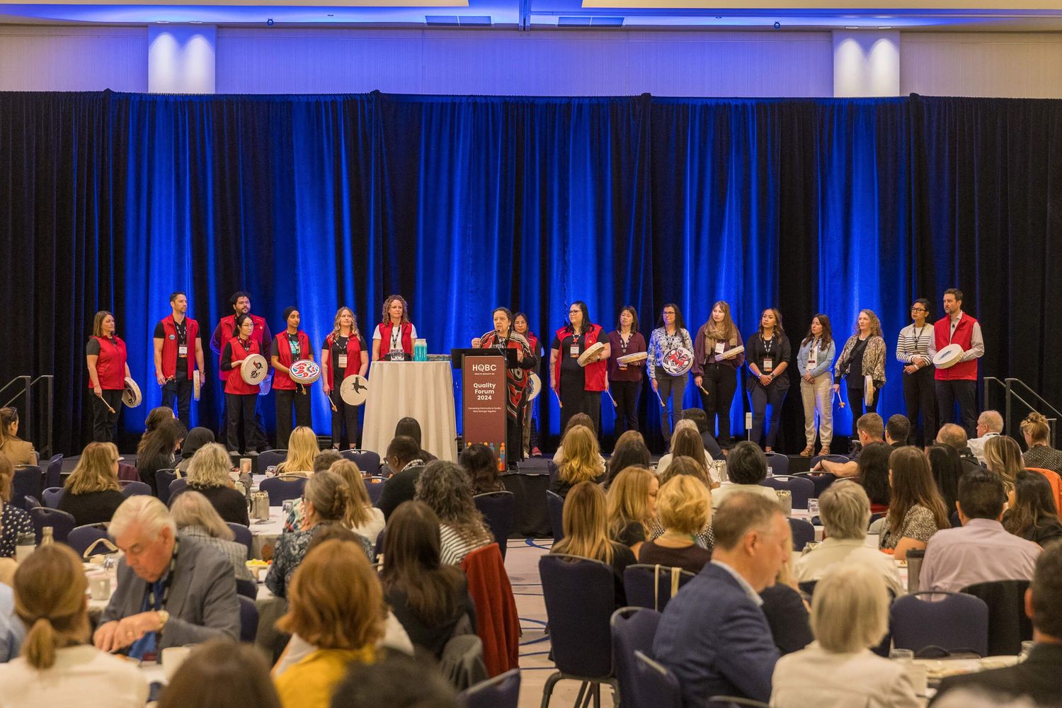 Group of women in red shirts on stage at a formal conference event with an audience seated in a ballroom.