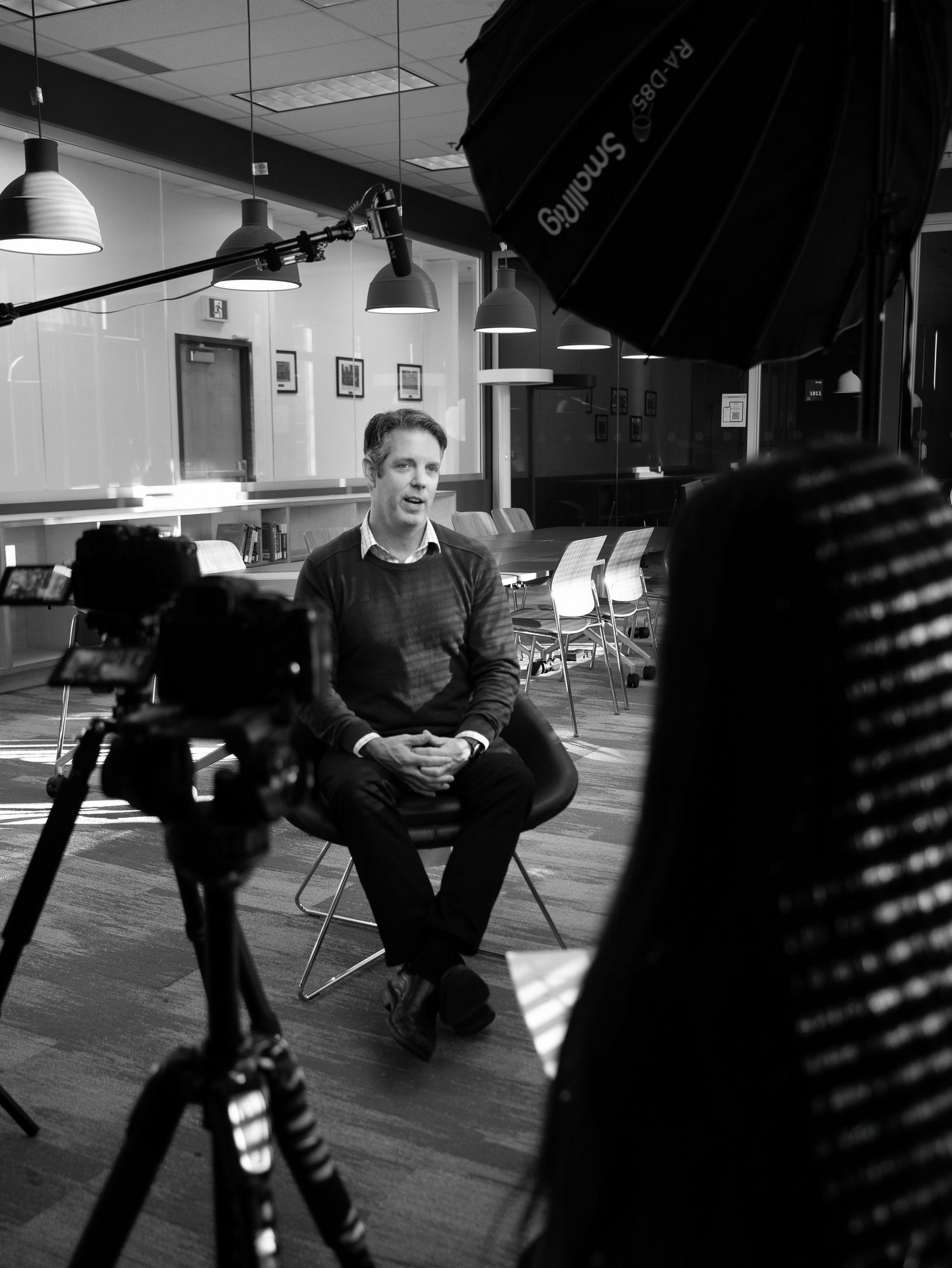 Black and white photo of a man being filmed in a professional video shoot setup with studio lights and camera equipment.