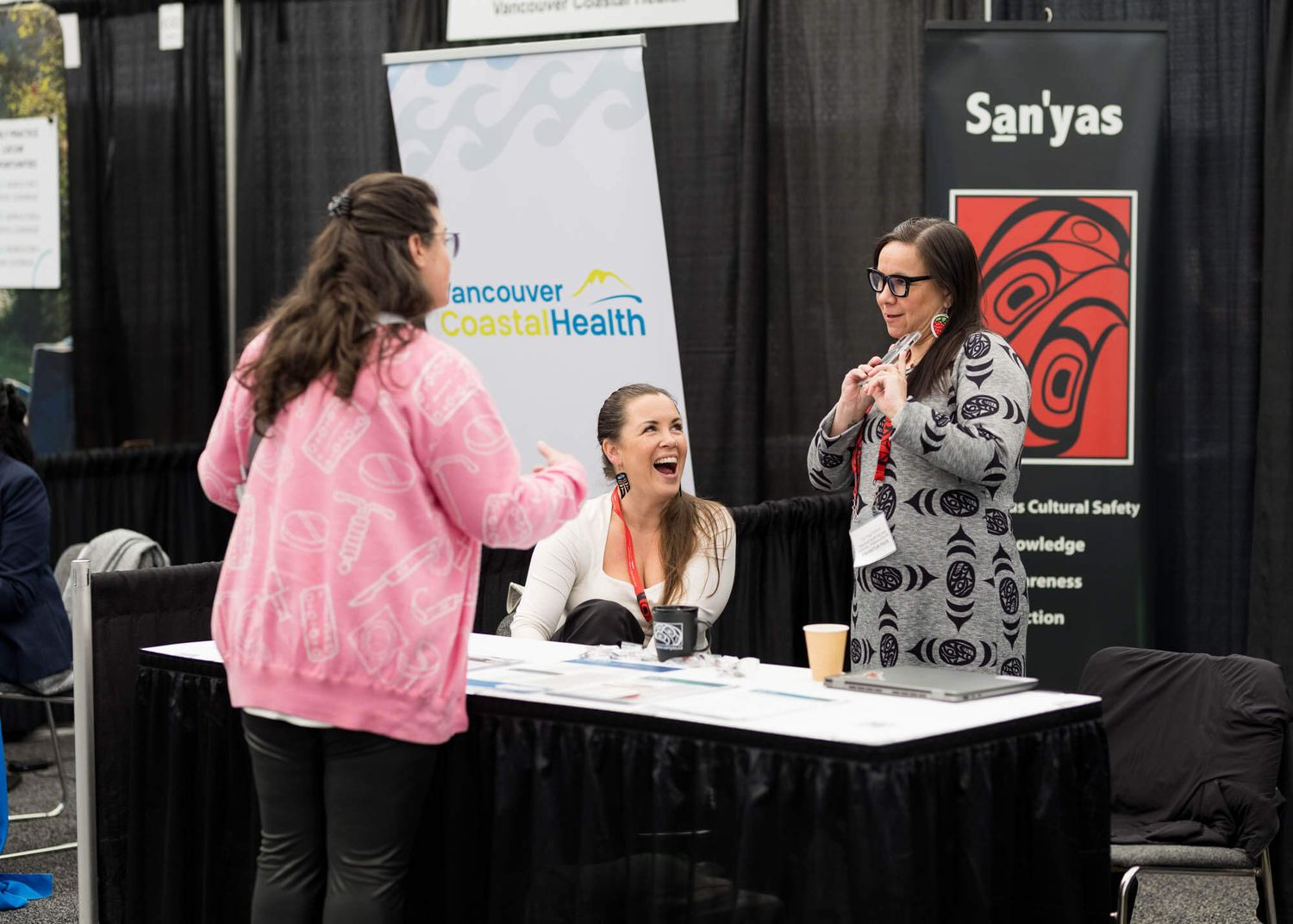 Three women networking at a Vancouver Coastal Health and San'yas booth during a health conference event.
