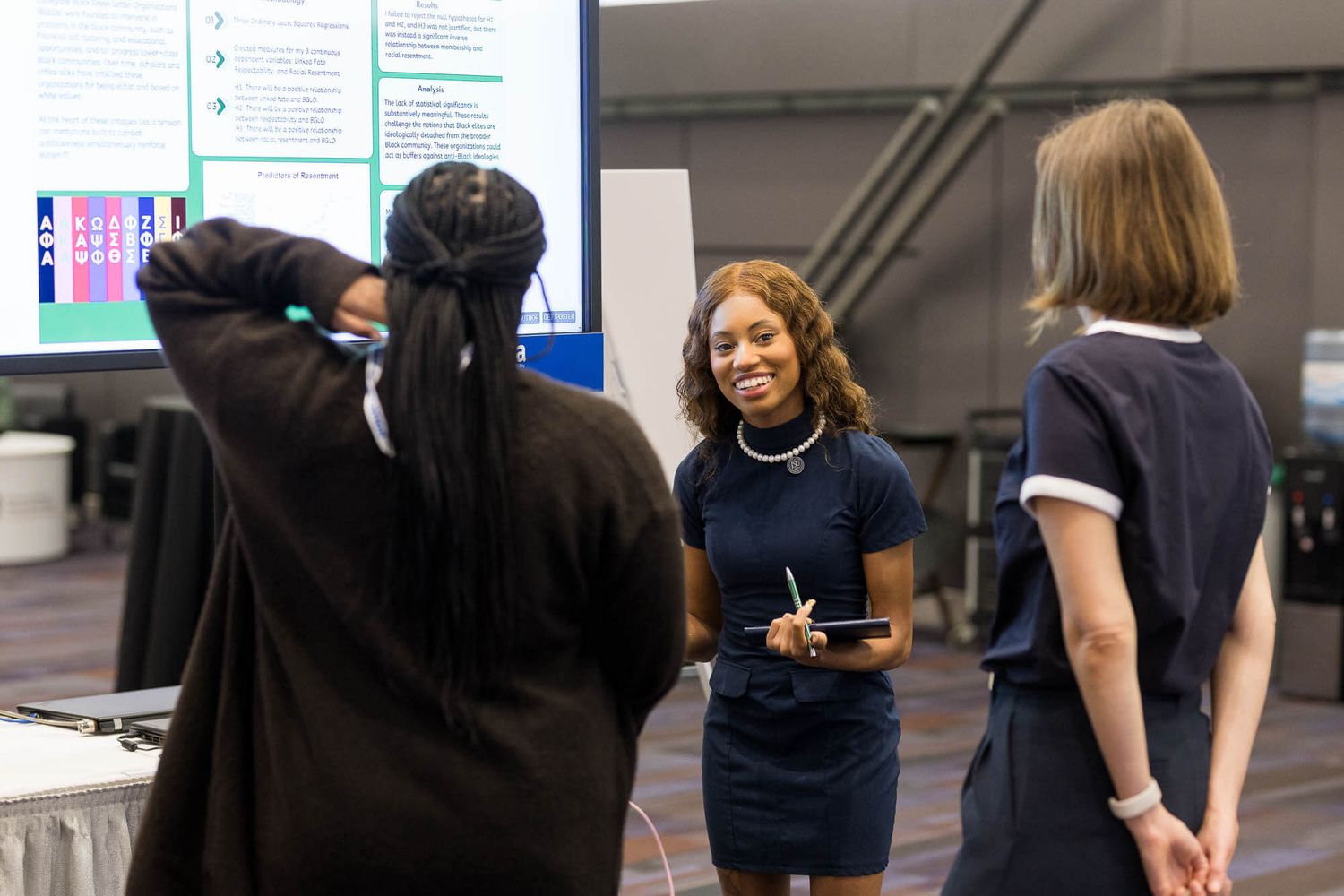 Three young women networking at a conference booth, smiling and discussing a research poster display.