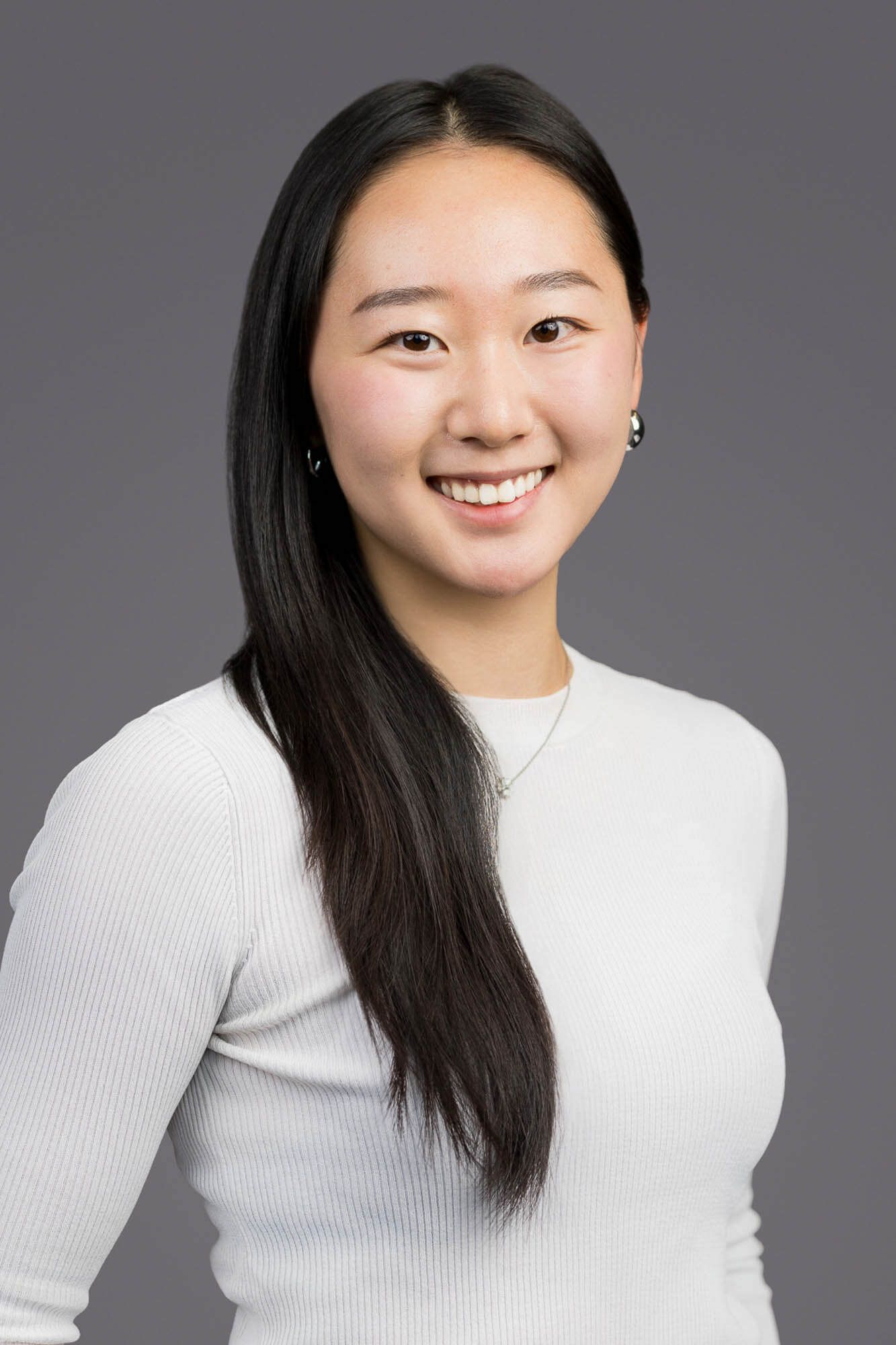 Professional headshot of a young woman with long black hair wearing a white ribbed top against a gray background.