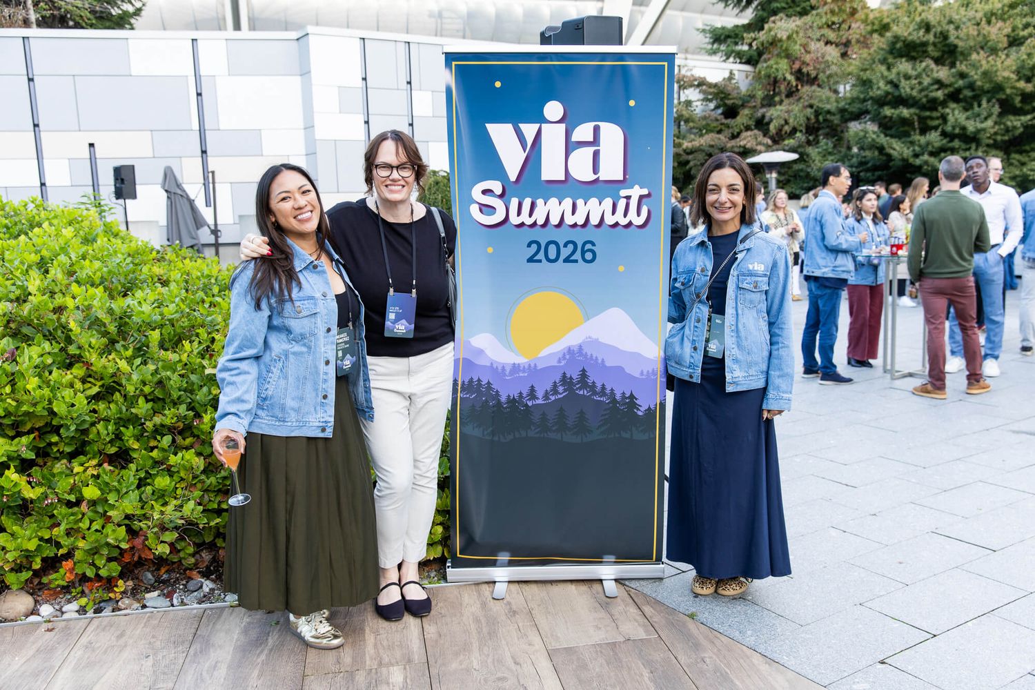 Three women pose together outdoors at the VIA Summit 2026 event, wearing matching denim jackets near a promotional banner.