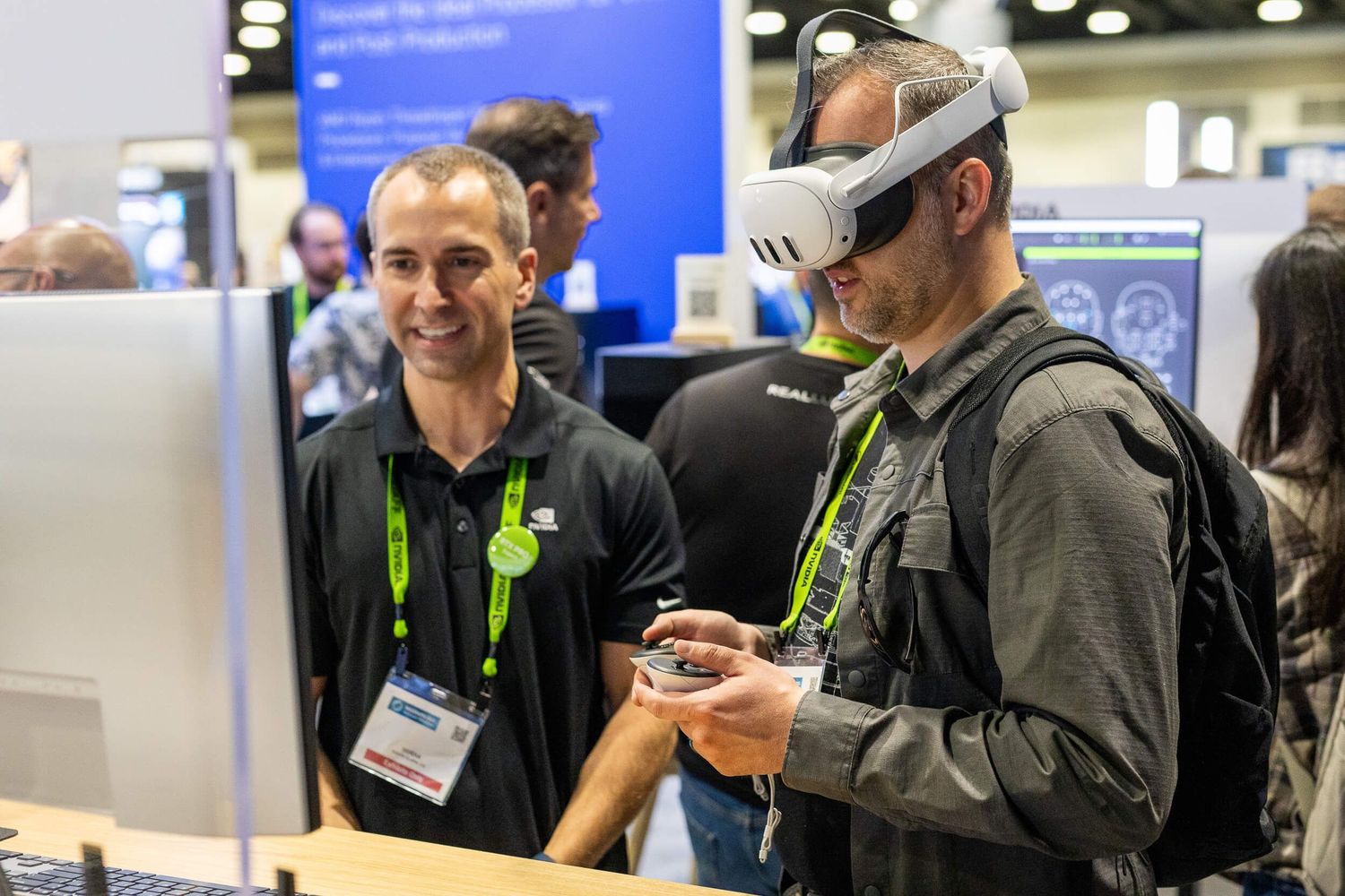 Two men at a tech expo, one wearing a VR headset and backpack, exploring virtual reality technology at a conference booth for Nvidia.