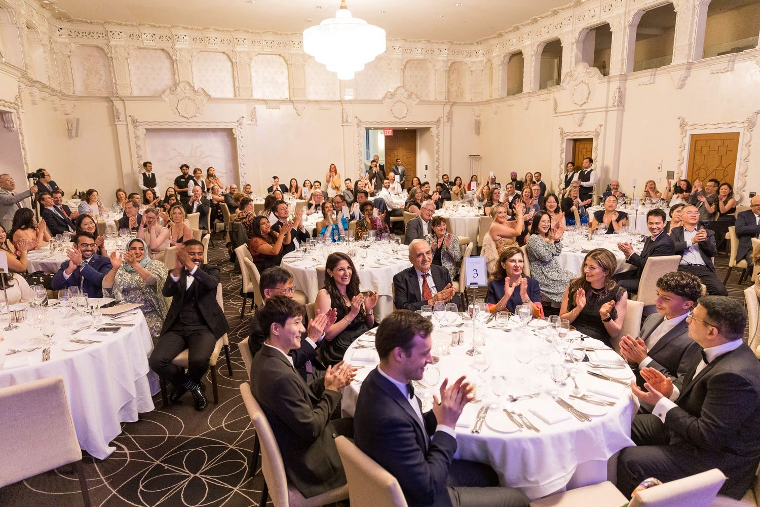 Guests applaud at an elegant formal gala dinner in a grand ballroom with round tables and chandeliers at the Hotel Georgia.