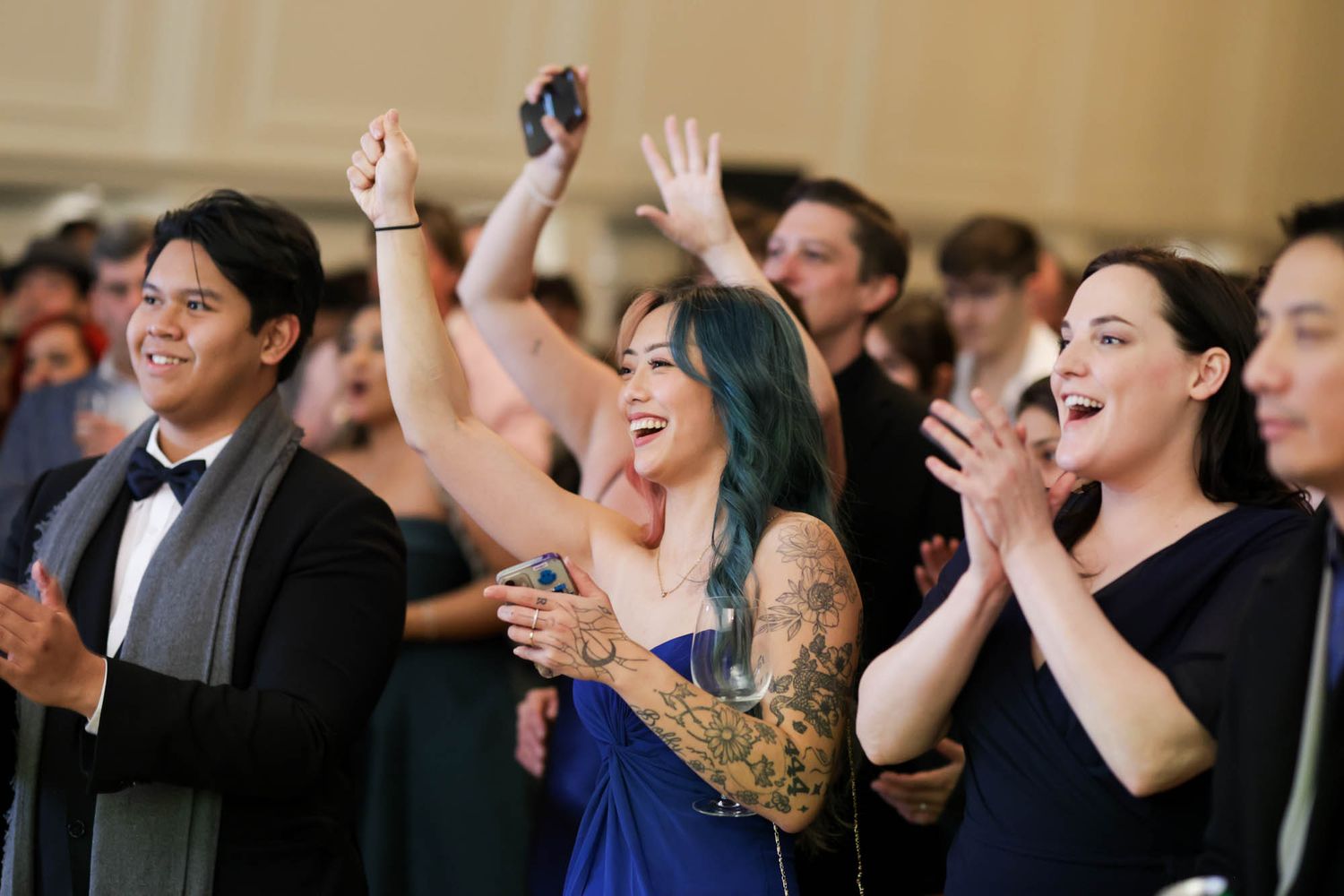 Excited guests cheering and clapping at an elegant formal event, woman with blue hair and tattoos raising hands in celebration.