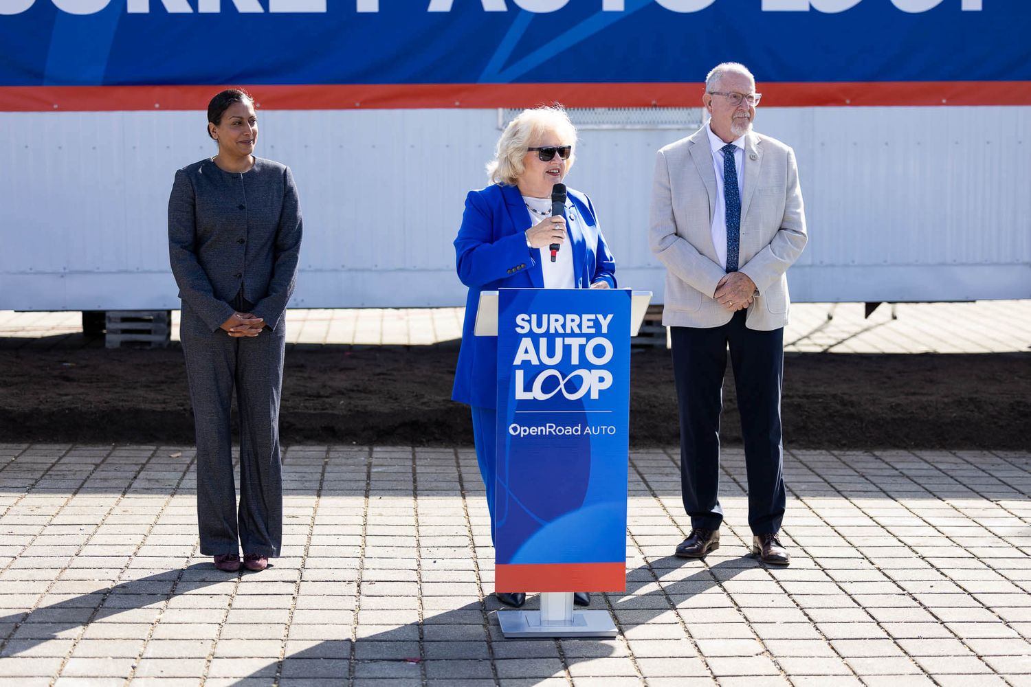 Woman in blue jacket speaks at Surrey Auto Loop podium flanked by two officials at outdoor press event.