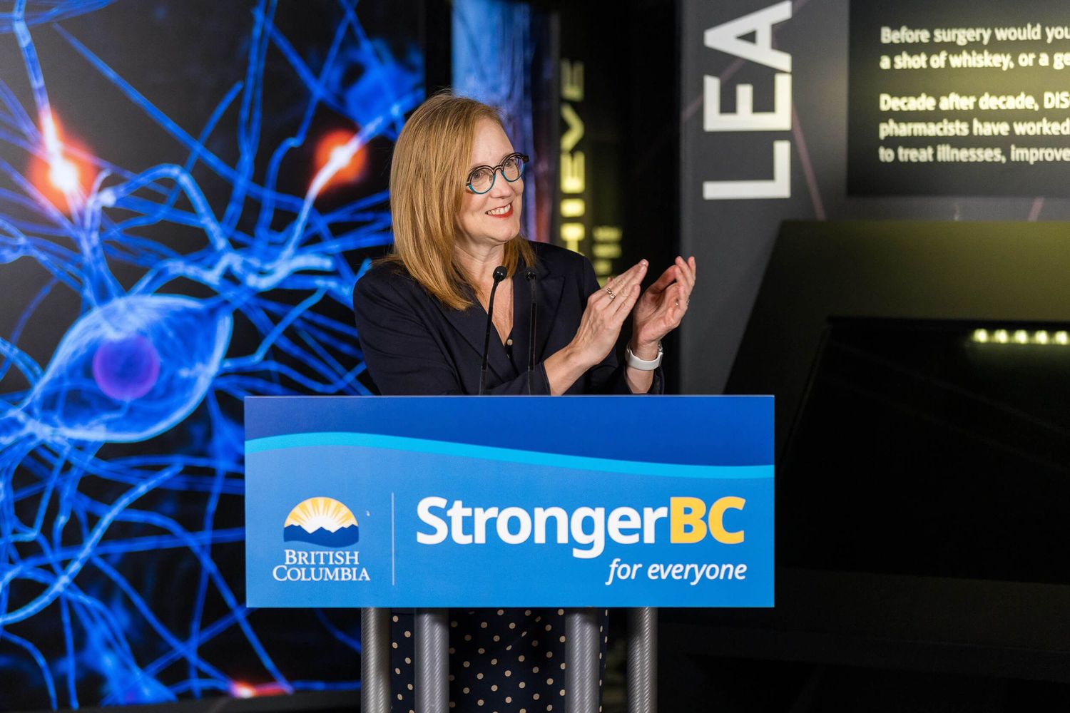 Brenda Bailey applauding at a StrongerBC for Everyone podium during a British Columbia government event.