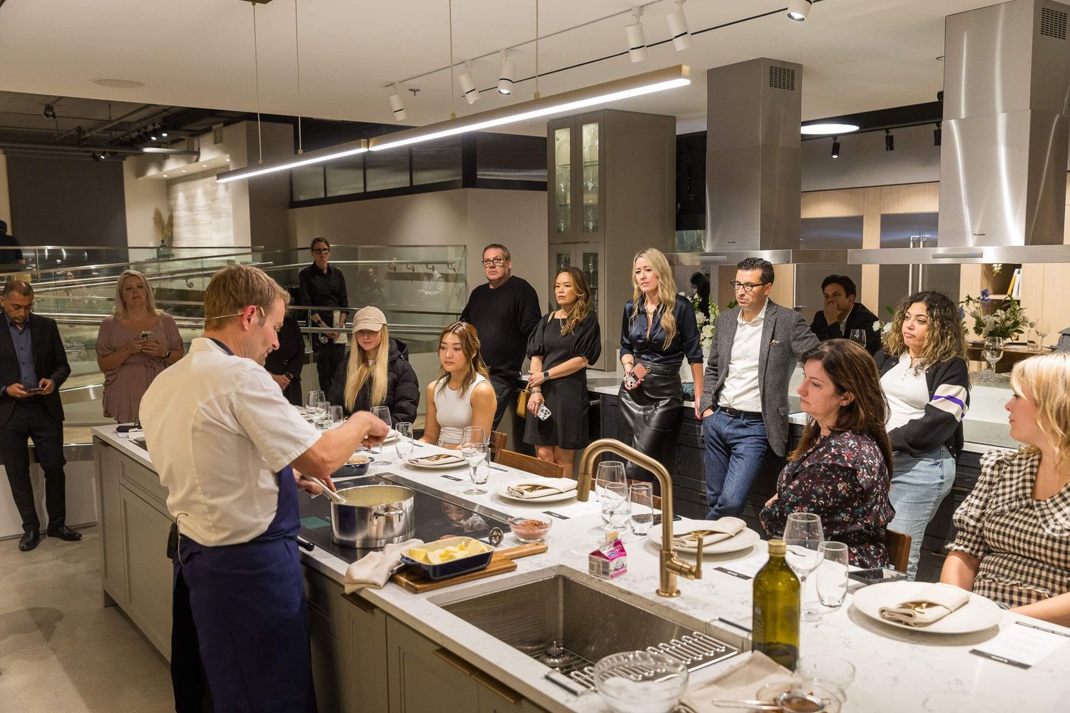 Chef demonstrates cooking techniques to guests gathered around a modern kitchen island in an upscale showroom setting for BOSCH Appliances.
