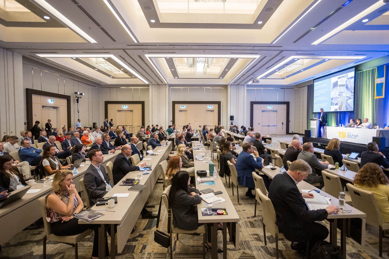 Large conference room filled with attendees seated at rows of tables during a professional business seminar event.