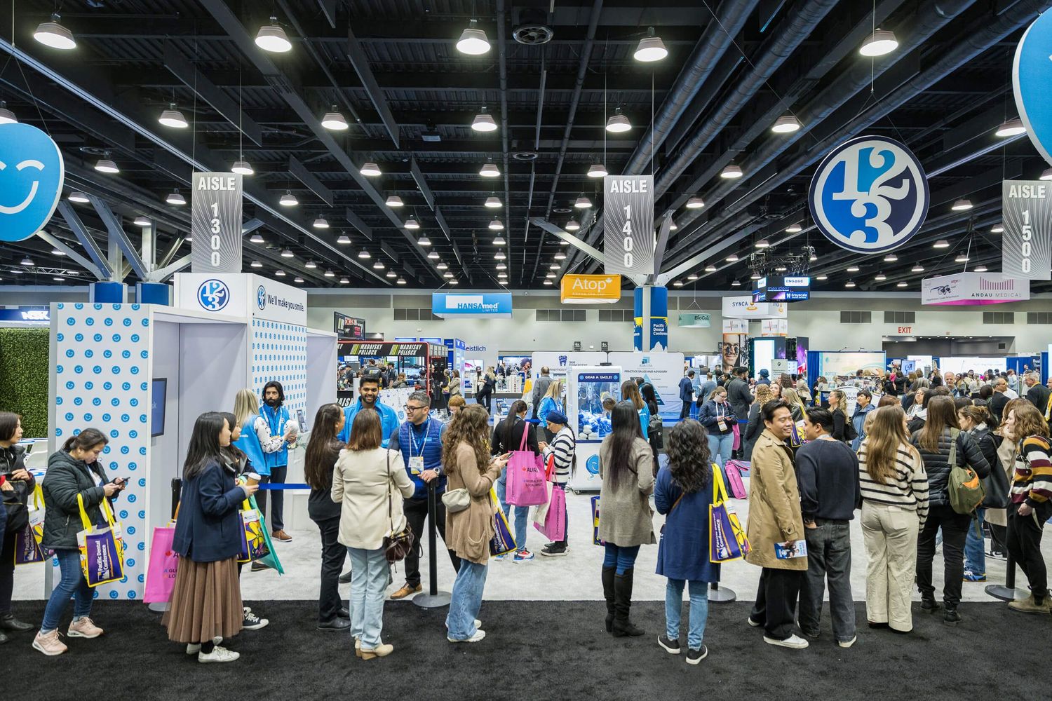 Crowded convention hall with exhibitor booths, attendees browsing displays under colorful branded signage at a trade show.