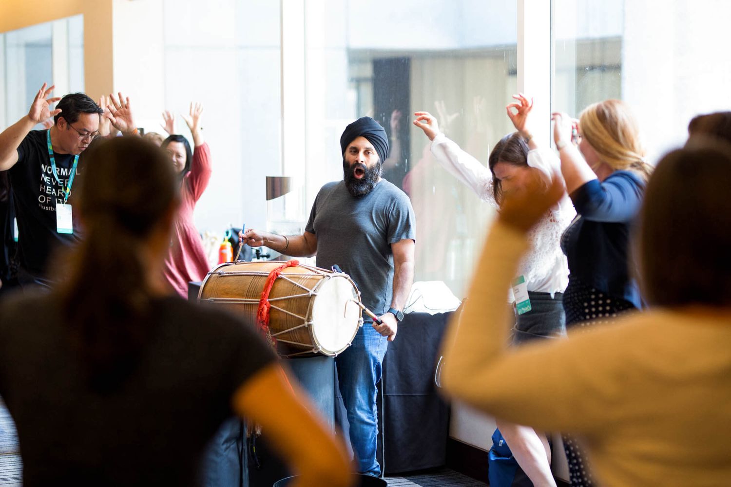 Man in blue turban plays dhol drum while diverse group of people dance with raised arms at cultural event.
