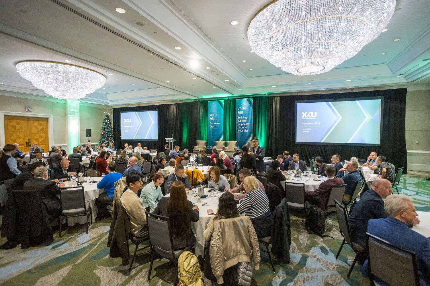Large conference banquet hall with attendees seated at round tables facing presentation screens at a formal business event.